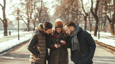 Three friends in warm winter coats and beanies standing together on a park path, looking intently at a smartphone held by a woman in the center.