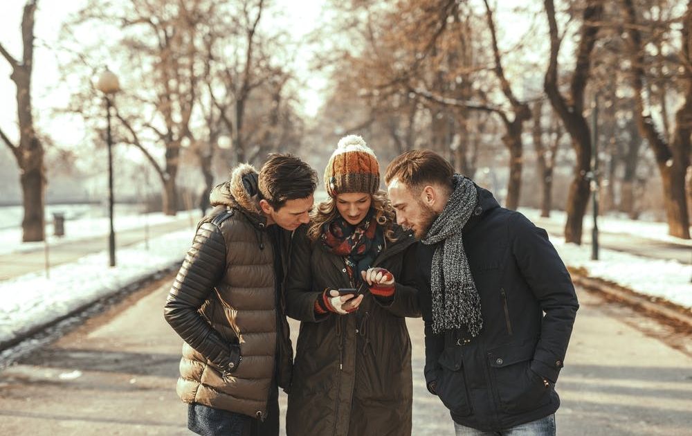 Three friends in warm winter coats and beanies standing together on a park path, looking intently at a smartphone held by a woman in the center.