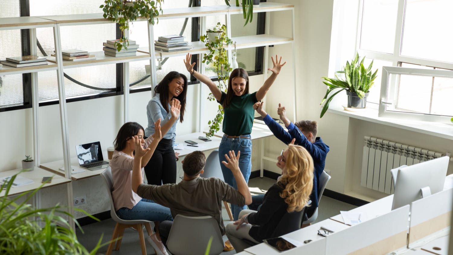 Bovenaanzicht van een creatief team van zes collega's die een succes vieren in een licht kantoor, met opgeheven handen, glimlachend en klappend samen.