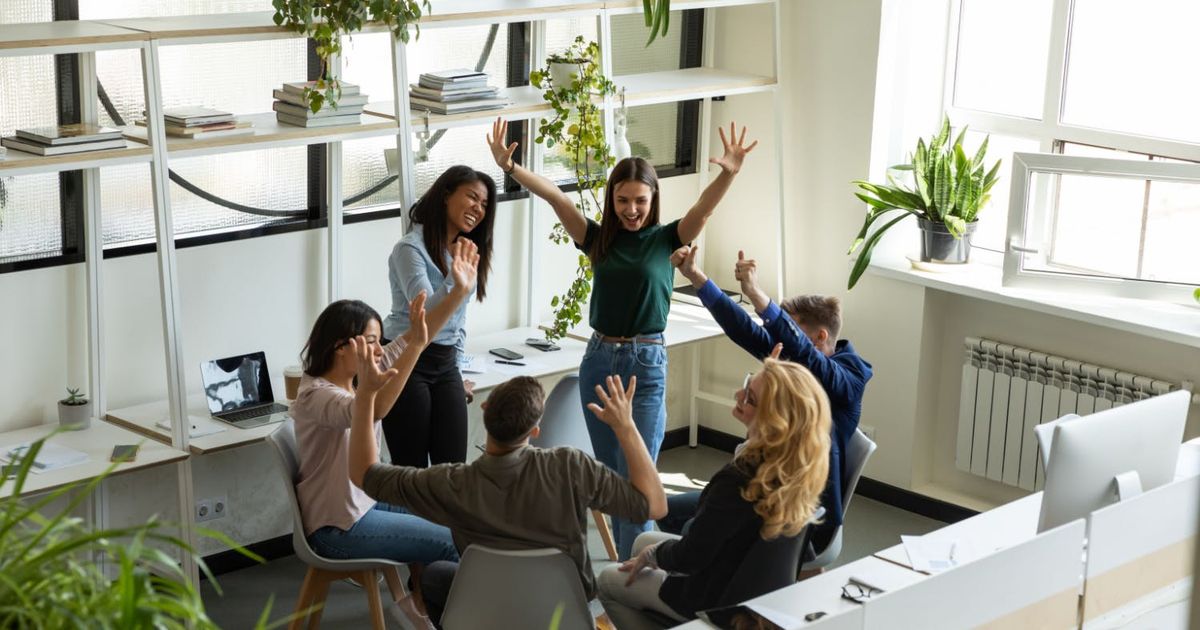 A high-angle view of a creative team of six colleagues celebrating a success in a bright office, with hands raised in the air, smiling and cheering together.