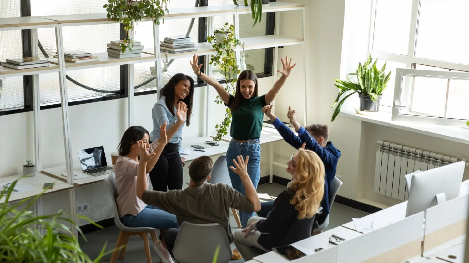 A high-angle view of a creative team of six colleagues celebrating a success in a bright office, with hands raised in the air, smiling and cheering together.
