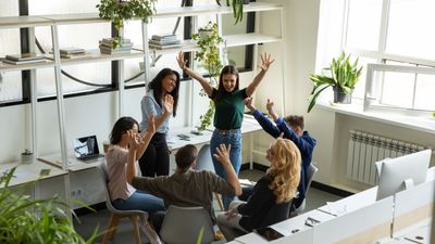 A high-angle view of a creative team of six colleagues celebrating a success in a bright office, with hands raised in the air, smiling and cheering together.