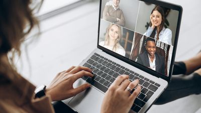 An over-the-shoulder view of a woman typing on her laptop while participating in a virtual meeting with four smiling colleagues displayed in a grid on her screen.