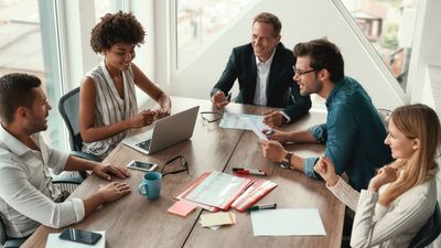 A high-angle view of a diverse team of five professionals smiling and collaborating around a wooden conference table during a bright office meeting.
