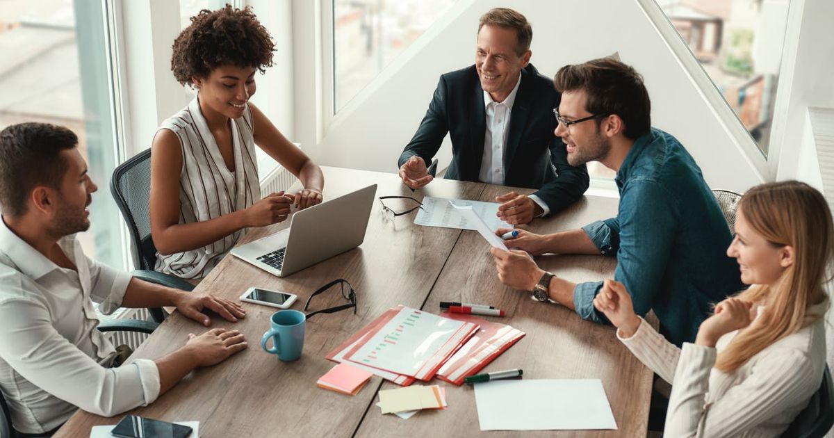 A high-angle view of a diverse team of five professionals smiling and collaborating around a wooden conference table during a bright office meeting.