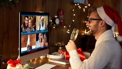 A smiling man in a Santa hat sits at his desk, raising a glass of champagne to toast his friends during a virtual Christmas celebration on his computer screen.