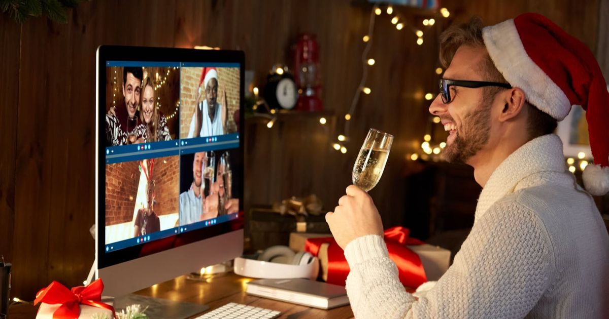 A smiling man in a Santa hat sits at his desk, raising a glass of champagne to toast his friends during a virtual Christmas celebration on his computer screen.