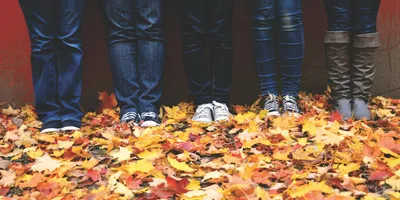 Four people standing side by side against a red wall in jeans, sneakers and boots, photographed from the knees down on a sidewalk thickly covered with yellow and orange fall leaves.